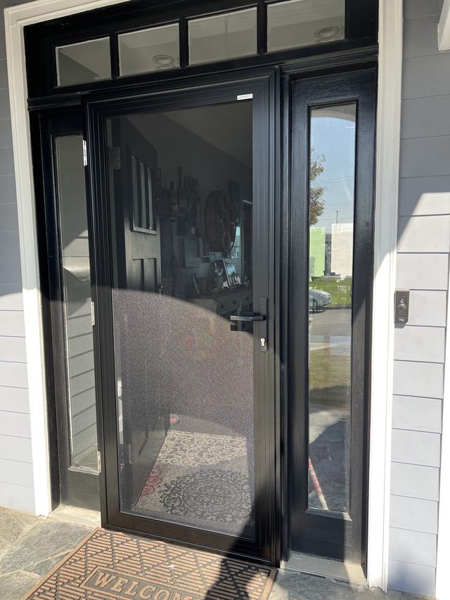 Black hinged security screen door on a front entry with a glass sidelight and transom windows, gray siding