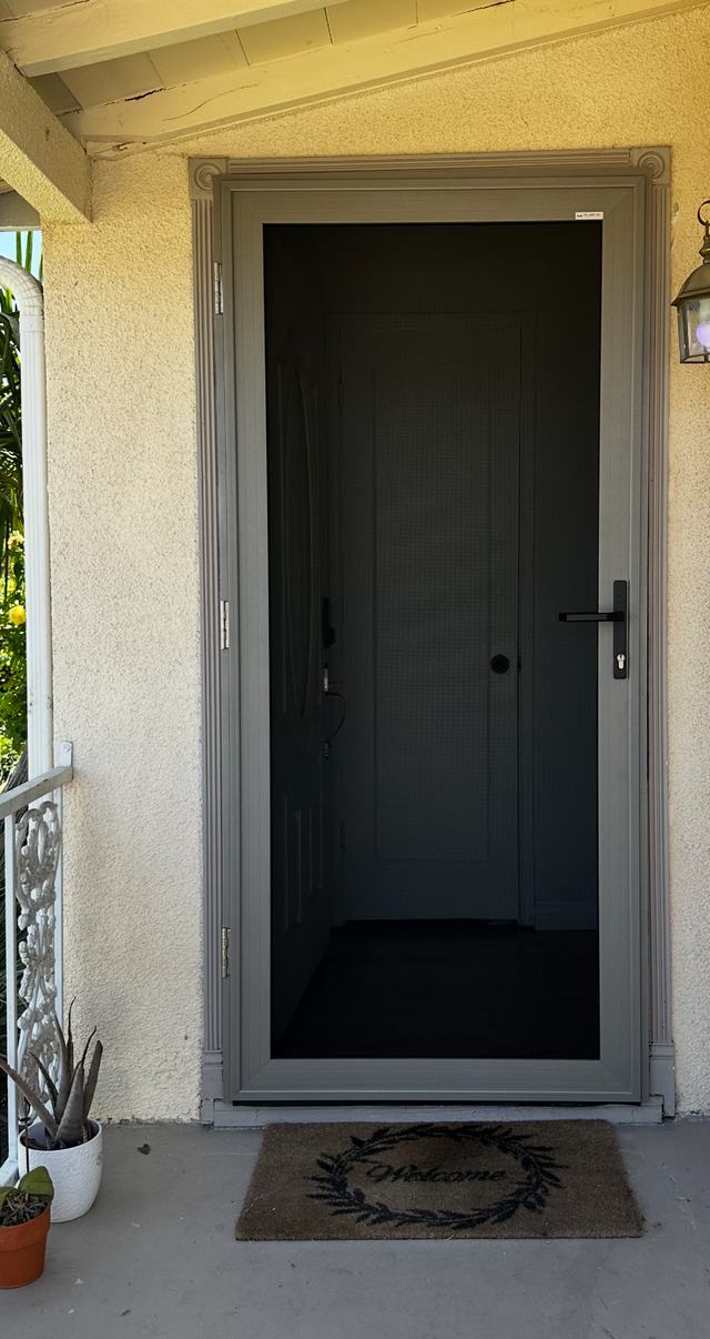 Gray hinged security screen door on a stucco front entry with a welcome mat