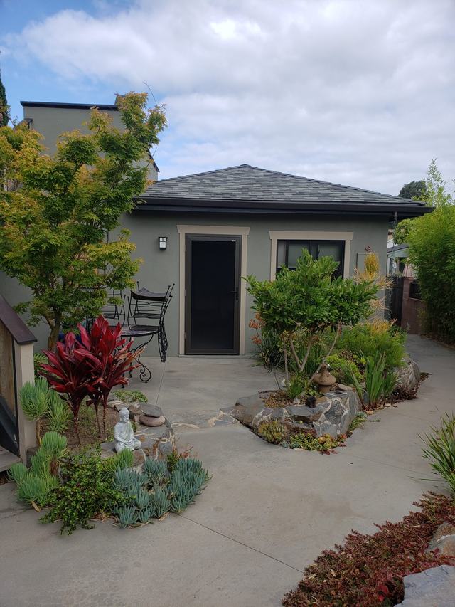 Black hinged security screen door on a small gray garden cottage behind a landscaped yard