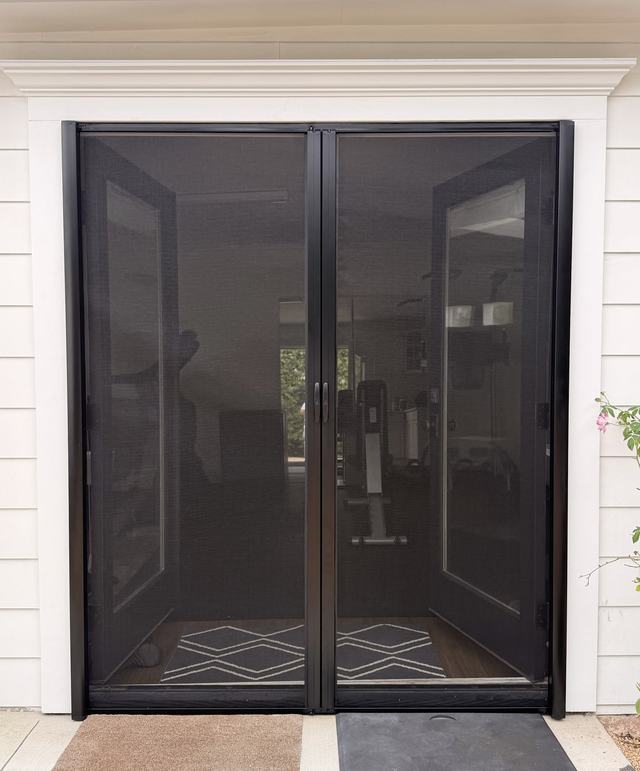 Black double retractable screen closed across open French doors on a white-sided house